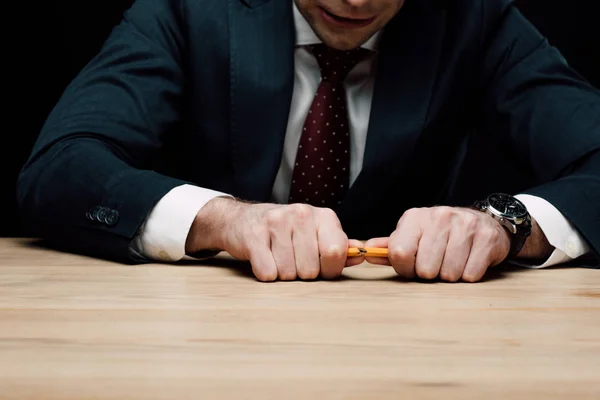 Angry businessman braking pencil while sitting at wooden desk isolated on black — Stock Photo