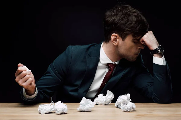 Depressed businessman sitting at desk near crumpled paper isolated on black — Stock Photo