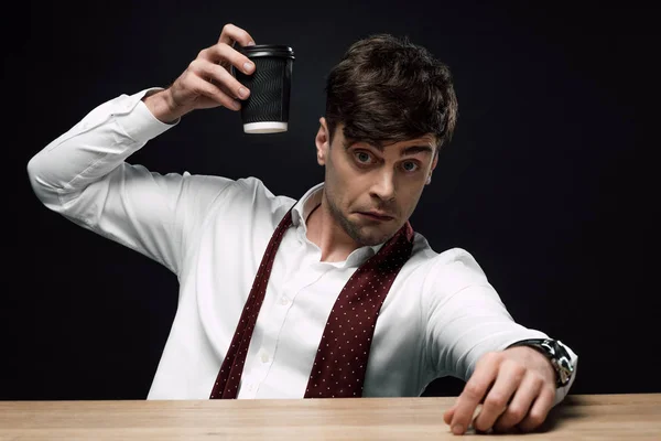 Hombre de negocios borracho levantando una taza de papel, mirando a la cámara y haciendo muecas aisladas en negro - foto de stock