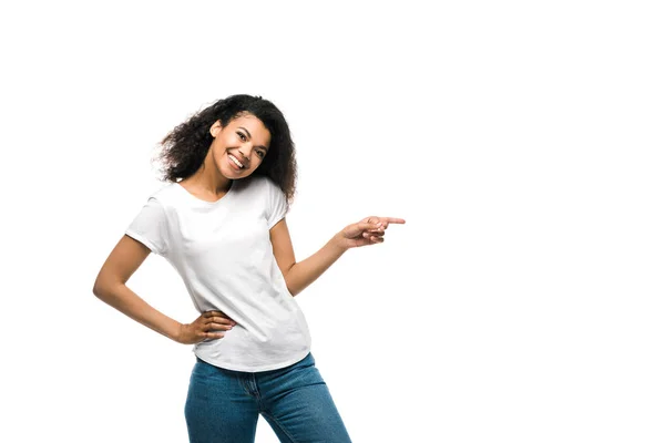 Menina americana africana feliz em t-shirt branca apontando com o dedo enquanto de pé com a mão no quadril isolado no branco — Fotografia de Stock