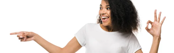 Tiro panorâmico de mulher americana africana alegre apontando com o dedo enquanto mostra sinal ok isolado no branco — Fotografia de Stock