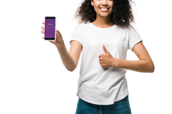 KYIV, UCRANIA - 29 de mayo de 2019: vista recortada de una chica estadounidense feliz sosteniendo un teléfono inteligente con aplicación instagram en la pantalla y mostrando el pulgar hacia arriba aislado en blanco - foto de stock
