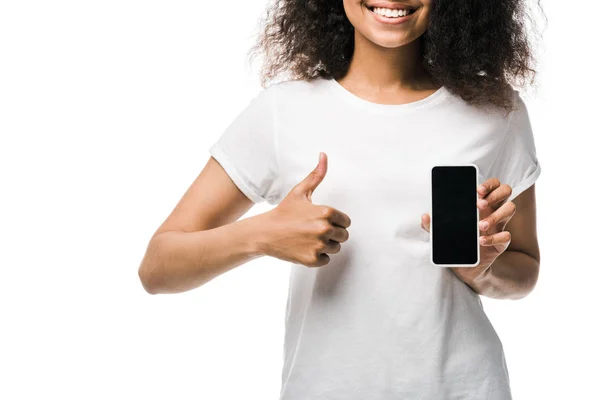 Обрізаний вид веселої американської дівчини, що тримає смартфон з порожнім екраном і показує великий палець вгору ізольовано на білому — стокове фото