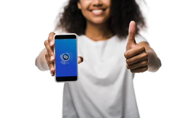 KYIV, UCRANIA - 29 de mayo de 2019: vista recortada de la niña afroamericana feliz sosteniendo el teléfono inteligente con la aplicación shazam en la pantalla y mostrando el pulgar hacia arriba aislado en blanco — Stock Photo