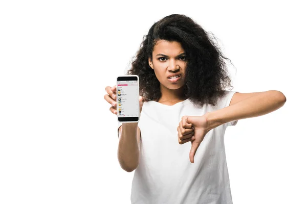 KYIV, UCRANIA - 29 de mayo de 2019: niña afroamericana disgustada sosteniendo un teléfono inteligente con música de manzana en la pantalla y mostrando el pulgar hacia abajo aislado en blanco - foto de stock