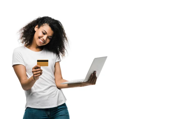 Alegre afroamericana mujer sosteniendo tarjeta de crédito cerca de portátil aislado en blanco - foto de stock
