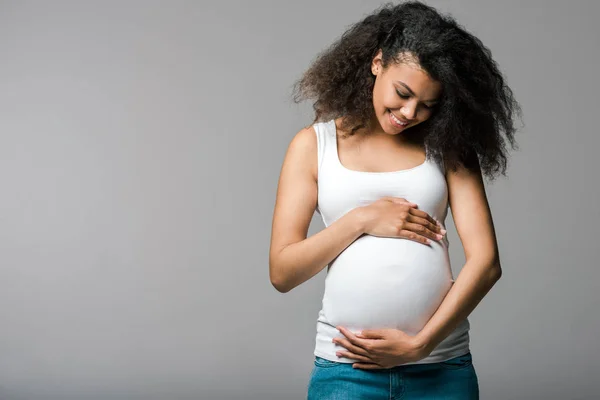 Alegre grávida afro-americana menina tocando barriga no cinza — Fotografia de Stock