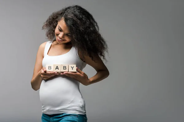 Fröhliche schwangere Afroamerikanerin hält Holzwürfel mit Baby-Schriftzug auf grau — Stockfoto