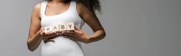 Plano panorámico de embarazada afroamericana chica sosteniendo cubos de madera con letras de bebé en gris - foto de stock
