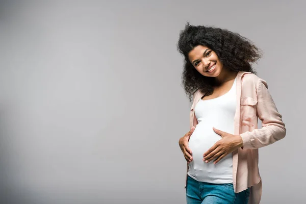 Positivo joven embarazada africano americano mujer tocando vientre en gris - foto de stock
