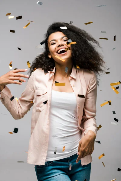 Happy african american girl smiling near shiny confetti and gesturing on grey — Stock Photo