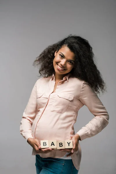 Chica afroamericana feliz sosteniendo cubos de madera con letras de bebé en gris - foto de stock