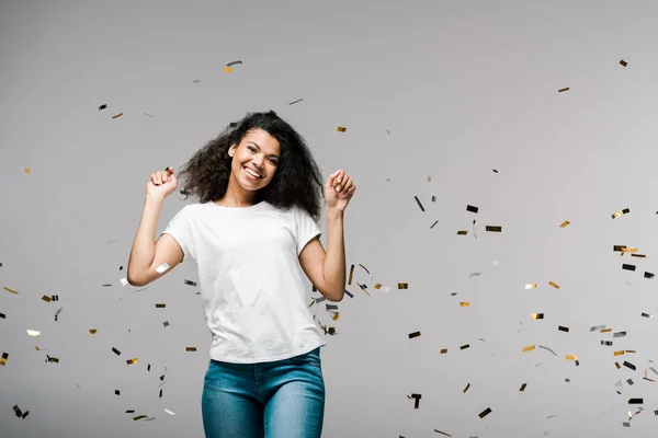 Cheerful african american girl smiling near shiny confetti while standing on grey — Stock Photo