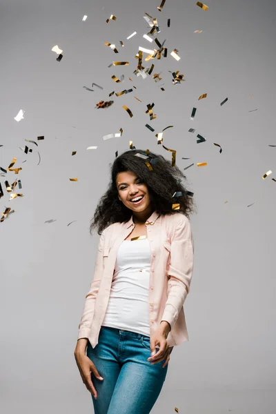 Young african american woman smiling near falling confetti while standing on grey — Stock Photo