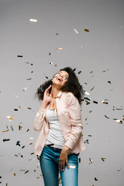 Happy african american woman smiling near falling confetti while standing on grey — Stock Photo