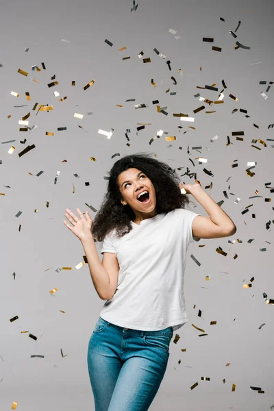 Young african american woman smiling and looking at falling confetti while standing on grey — Stock Photo