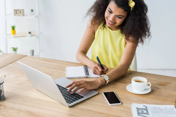 Glückliche afrikanisch-amerikanische Frau hält Stift in der Nähe von Notizbuch und schaut auf Laptop — Stockfoto