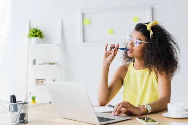 Nachdenkliche junge afrikanisch-amerikanische Frau mit Brille hält Stift in der Hand, während sie neben Laptop sitzt — Stockfoto