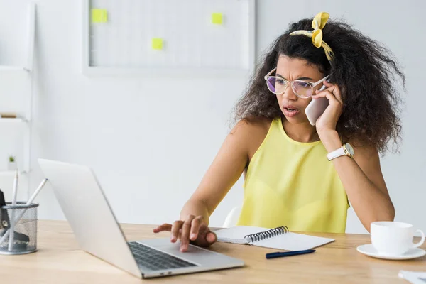 Atractiva mujer afroamericana en gafas usando el ordenador portátil mientras habla en el teléfono inteligente - foto de stock