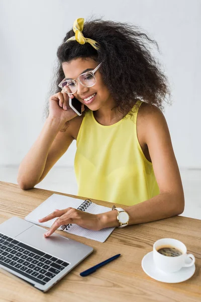 Happy african american woman in glasses using laptop while talking on smartphone near cup of coffee — Stock Photo