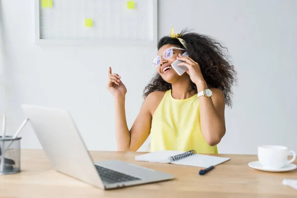 Glückliche junge afrikanisch-amerikanische Frau mit Brille gestikuliert und benutzt Laptop, während sie auf dem Smartphone spricht — Stockfoto