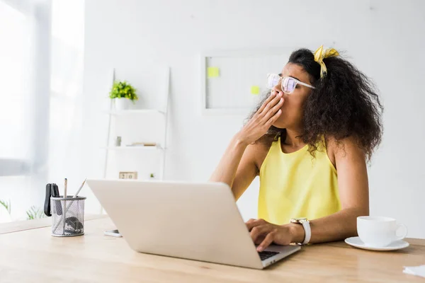 Femme afro-américaine fatiguée dans des lunettes bâillant et couvrant la bouche près d'un ordinateur portable — Photo de stock