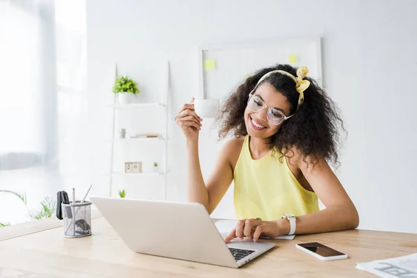 Щаслива афроамериканська бізнес-леді в окулярах дивиться на ноутбук, тримаючи чашку — стокове фото