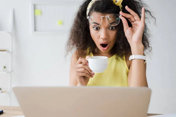 Enfoque selectivo de impactada chica afroamericana tocando gafas mientras mira el ordenador portátil y la celebración de la taza de café - foto de stock