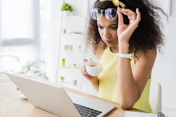 Disgustado afroamericano chica tocando gafas mientras mira el ordenador portátil y la celebración de la taza de café - foto de stock