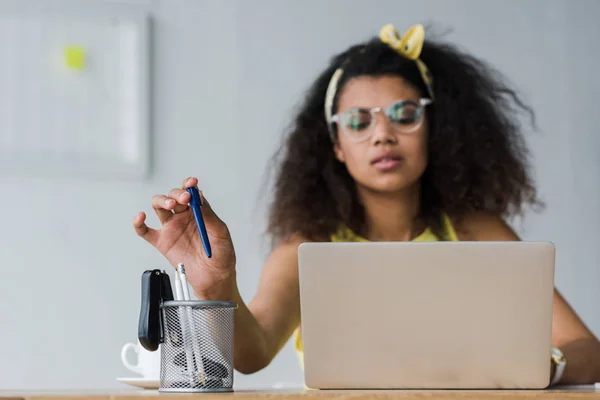 Enfoque selectivo de titular de la pluma y la mujer afroamericana tomando pluma mientras se utiliza el ordenador portátil - foto de stock