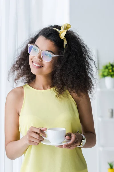 Fröhliche afrikanisch-amerikanische Frau in Gläsern mit Untertasse und Tasse mit Getränk — Stockfoto