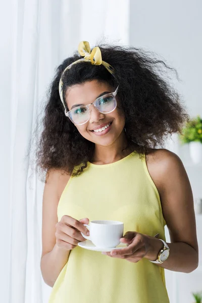 Heureuse femme afro-américaine dans des lunettes tenant soucoupe et tasse avec boisson — Photo de stock