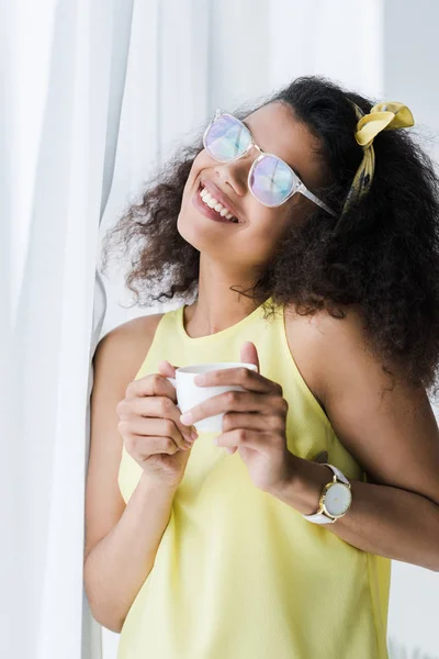 Mujer afroamericana feliz en vasos sosteniendo taza con bebida - foto de stock