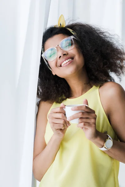 Mujer afroamericana feliz en vasos sosteniendo taza con bebida - foto de stock