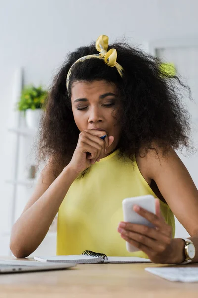 Selektiver Fokus einer müden afrikanisch-amerikanischen Frau, die gähnt und ihr Gesicht verdeckt, während sie auf ihr Smartphone schaut — Stockfoto