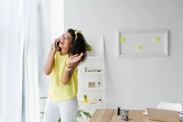 Feliz rizado africano americano mujer hablando en teléfono inteligente cerca del ordenador portátil - foto de stock