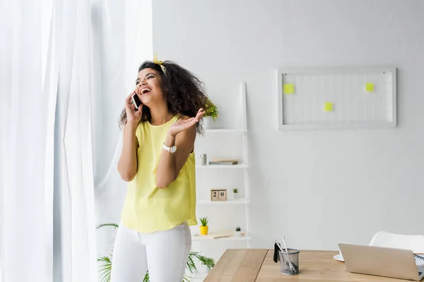 Alegre rizado africano americano mujer hablando en teléfono inteligente cerca del ordenador portátil - foto de stock