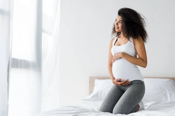 Alegre africano americano embarazada mujer tocando vientre mientras sentado en cama - foto de stock