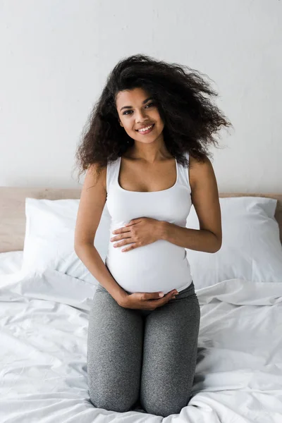 Счастливая кудрявая африканская беременная американка, прикасающаяся к животу сидя на кровати — стоковое фото