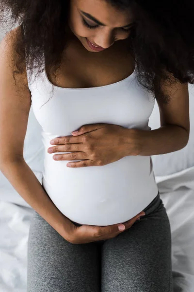 Overhead-Ansicht von positiven lockigen afrikanisch-amerikanischen schwangeren Frau, die Bauch berühren, während sie auf dem Bett sitzt — Stockfoto