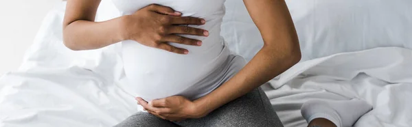 Panoramic shot of pregnant african american woman touching belly while sitting on bed — Stock Photo
