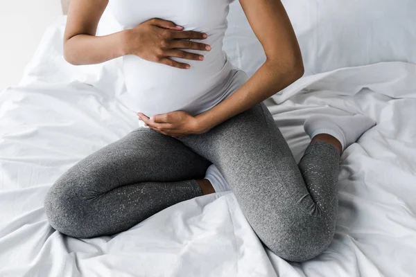 Vista recortada de la mujer afroamericana embarazada tocando el vientre mientras está sentado en la cama - foto de stock