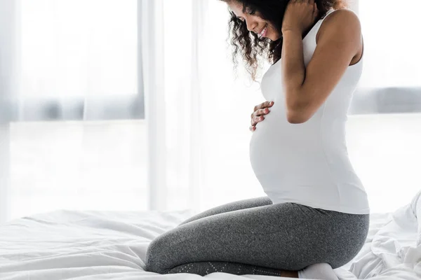 Cropped view of happy curly pregnant african american woman touching belly while sitting on bed — Stock Photo