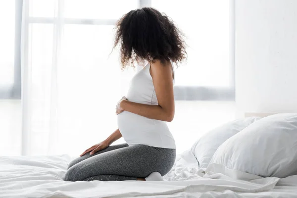 Беременная американка из Африки трогает живот, сидя на кровати — стоковое фото