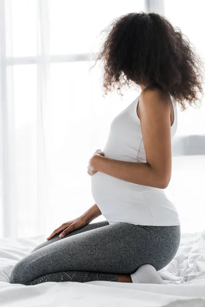 Morena embarazada africana americana mujer tocando vientre mientras sentado en la cama - foto de stock