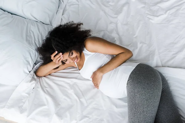 Vista dall'alto della felice donna africana americana incinta che tocca la pancia mentre giace sul letto — Foto stock