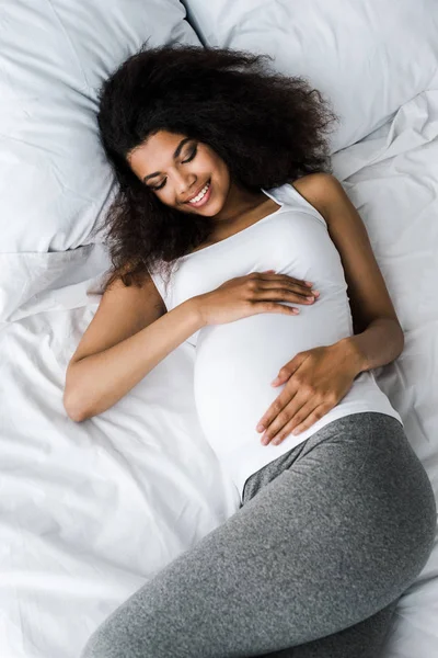 Вид сверху на улыбающуюся беременную африканскую женщину лежащую на кровати — стоковое фото