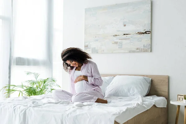 Attractive pregnant african american woman looking at belly and sitting on bed — Stock Photo