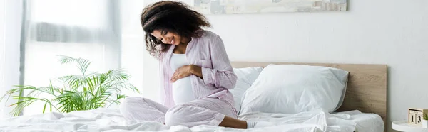 Panoramic shot of attractive pregnant african american woman looking at belly and sitting on bed — Stock Photo