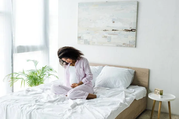 Beautiful young pregnant african american woman looking at belly and sitting on bed — Stock Photo